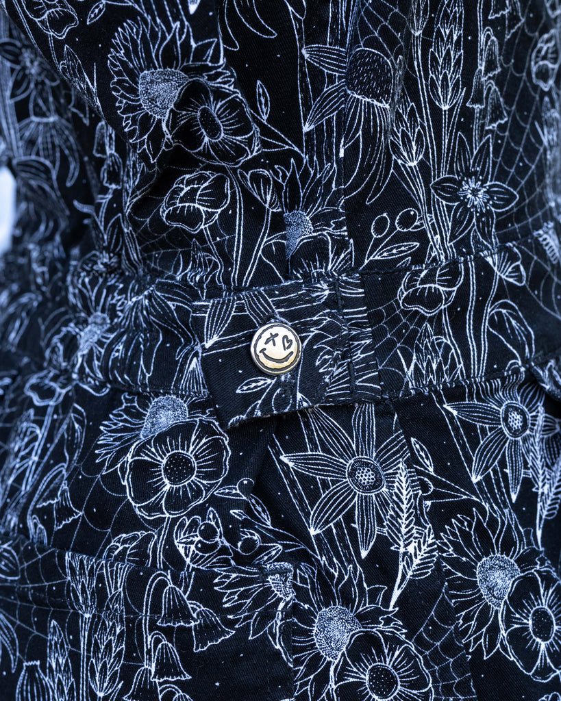 Close-up of the embroidered Rebel Romance logo button on the Black Floral Cobweb Boilersuit. The black stretch twill fabric features an intricate white cobweb and floral pattern.
