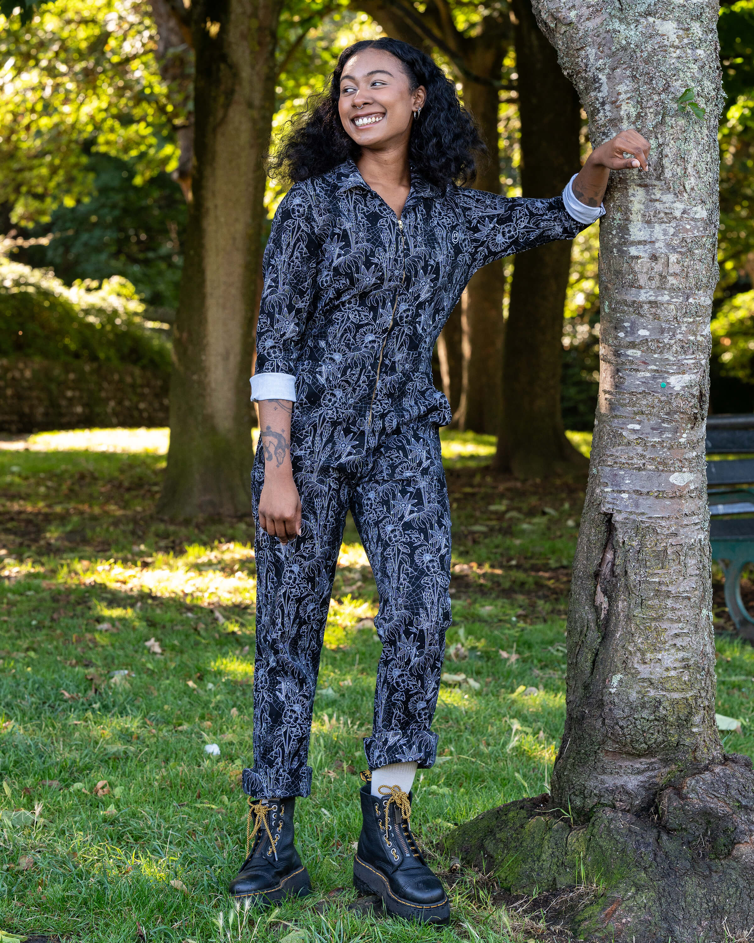 Jasmine leans on a tree wearing the Rebel Romance Black Floral Cobweb Boilersuit. The alternative one-piece features long sleeves, straight legs, and a zip fastening in black with a white floral cobweb print.