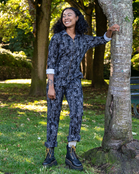 Jasmine leans on a tree wearing the Rebel Romance Black Floral Cobweb Boilersuit. The alternative one-piece features long sleeves, straight legs, and a zip fastening in black with a white floral cobweb print.