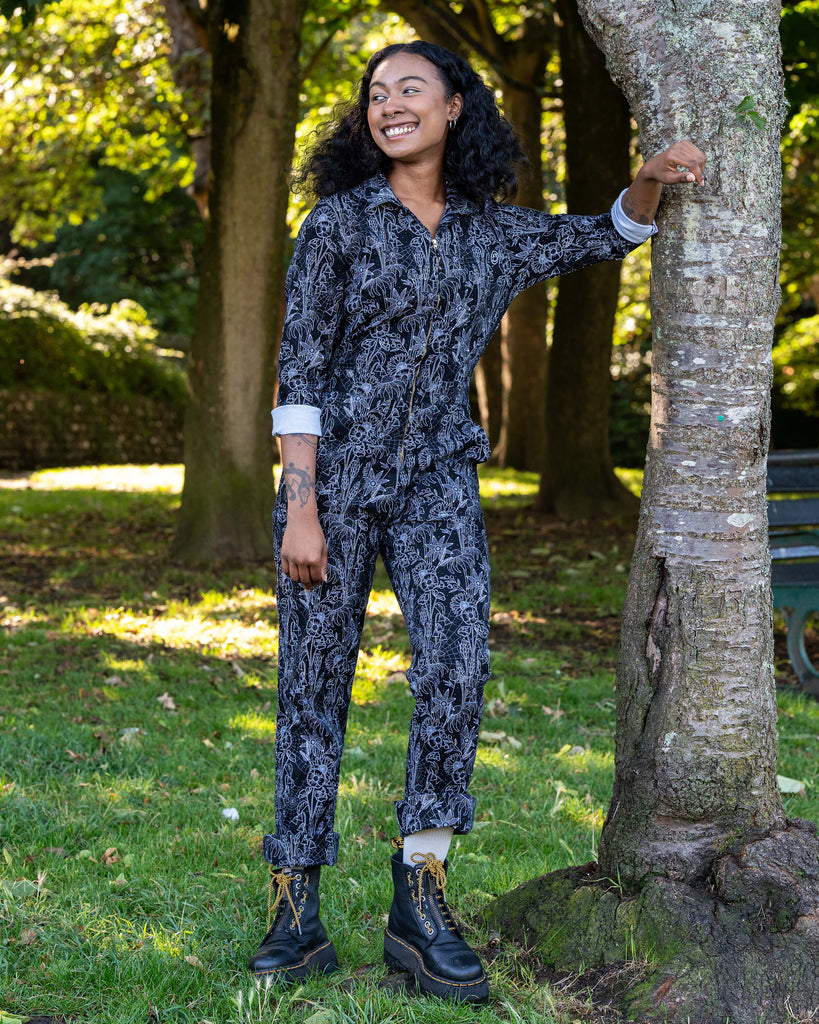 Jasmine leans on a tree wearing the Rebel Romance Black Floral Cobweb Boilersuit. The alternative one-piece features long sleeves, straight legs, and a zip fastening in black with a white floral cobweb print.