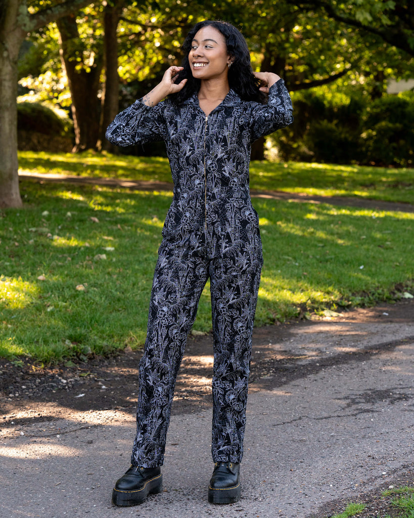 Jasmine wears the Rebel Romance Black Floral Cobweb Boilersuit in a park setting. The black one-piece features a white floral and cobweb print, a zip front, long sleeves, and straight legs. Ethically made and designed for comfort and alternative style.
