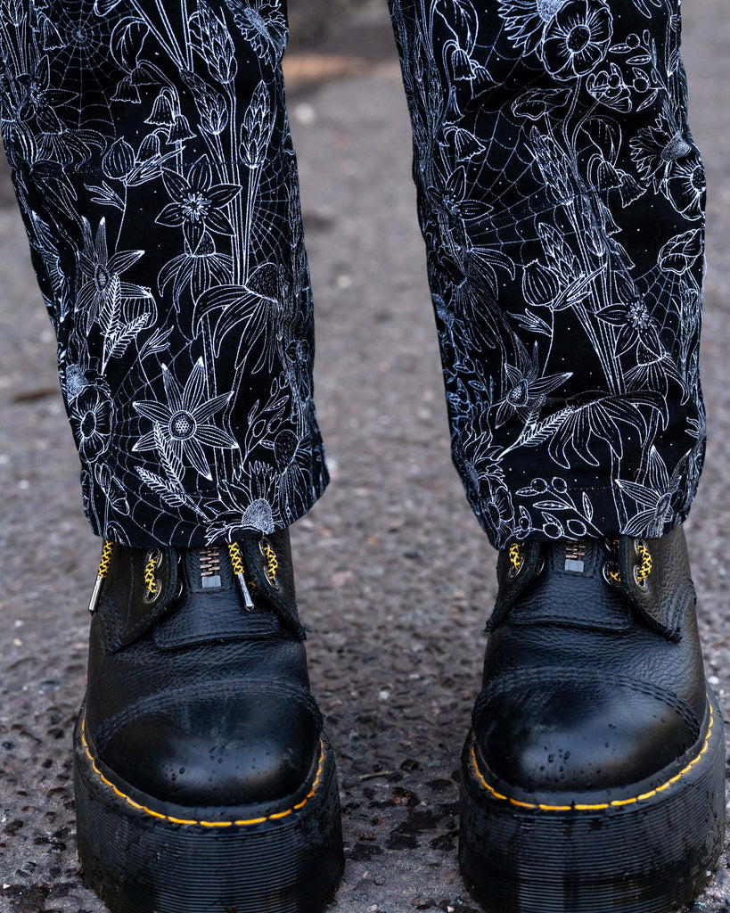 Jasmine wears the Rebel Romance Black Floral Cobweb Boilersuit, showing the back design. The all-over floral and cobweb print covers the black stretch twill fabric. Ethical and inclusive slow fashion brand.