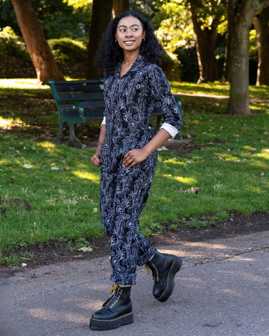 Jasmine poses side-on wearing the Rebel Romance Black Floral Cobweb Boilersuit. The one-piece features long sleeves, pockets, and a straight-leg fit in black twill with a white floral cobweb print.