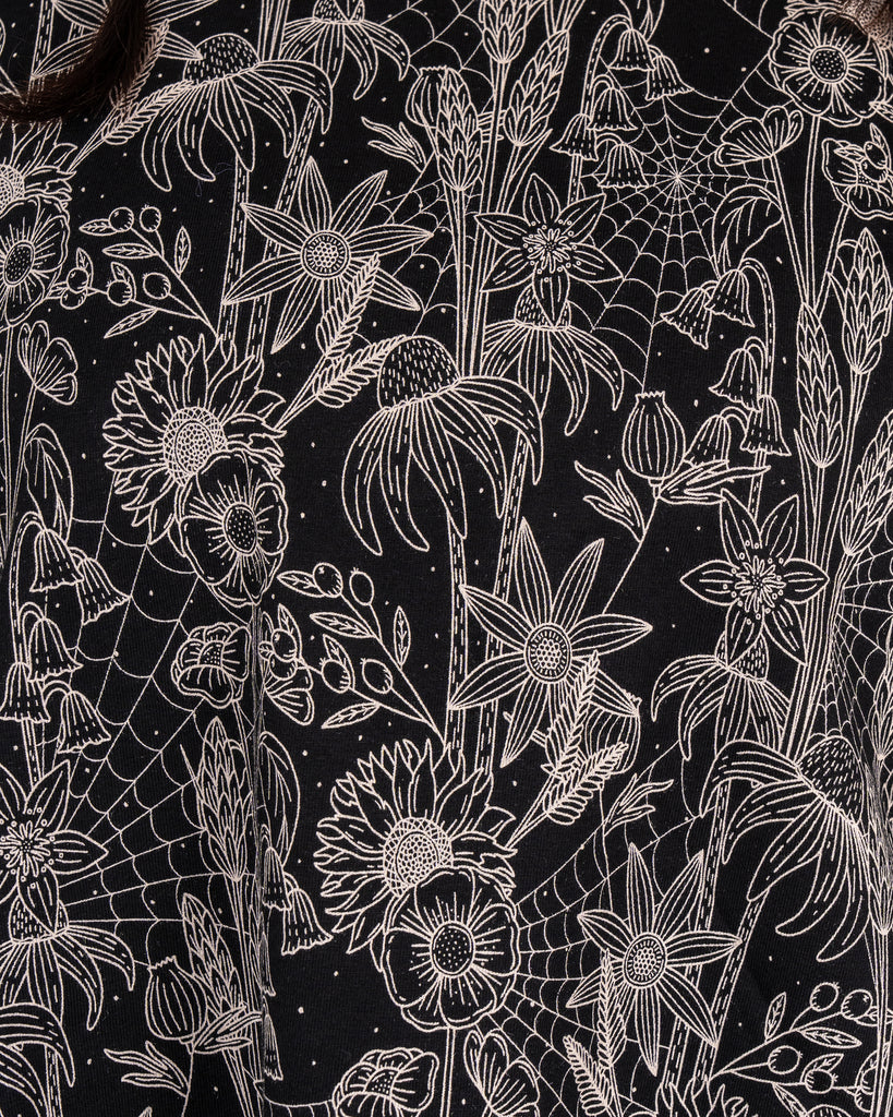 Macro close-up of the Rebel Romance Black Floral Cobweb Cropped Boxy Tee print, featuring detailed beige flowers, grasses and spiderwebs on black. This bold botanical print is part of Rebel Romance’s alternative slow fashion range made in the UK.