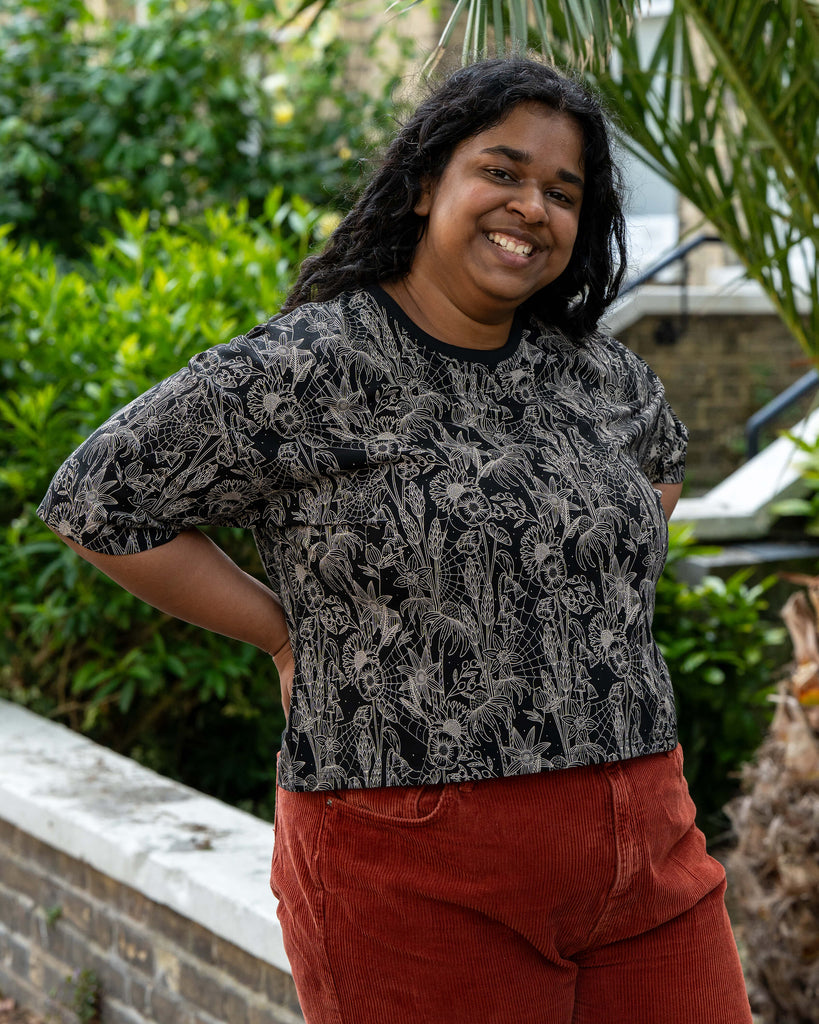 Vidhya poses casually in the Rebel Romance Black Floral Cobweb Cropped Boxy Tee. The cropped tee features a magical beige botanical print with spiderwebs, paired with rust red corduroy trousers. Rebel Romance embraces alternative fashion for UK millennials.