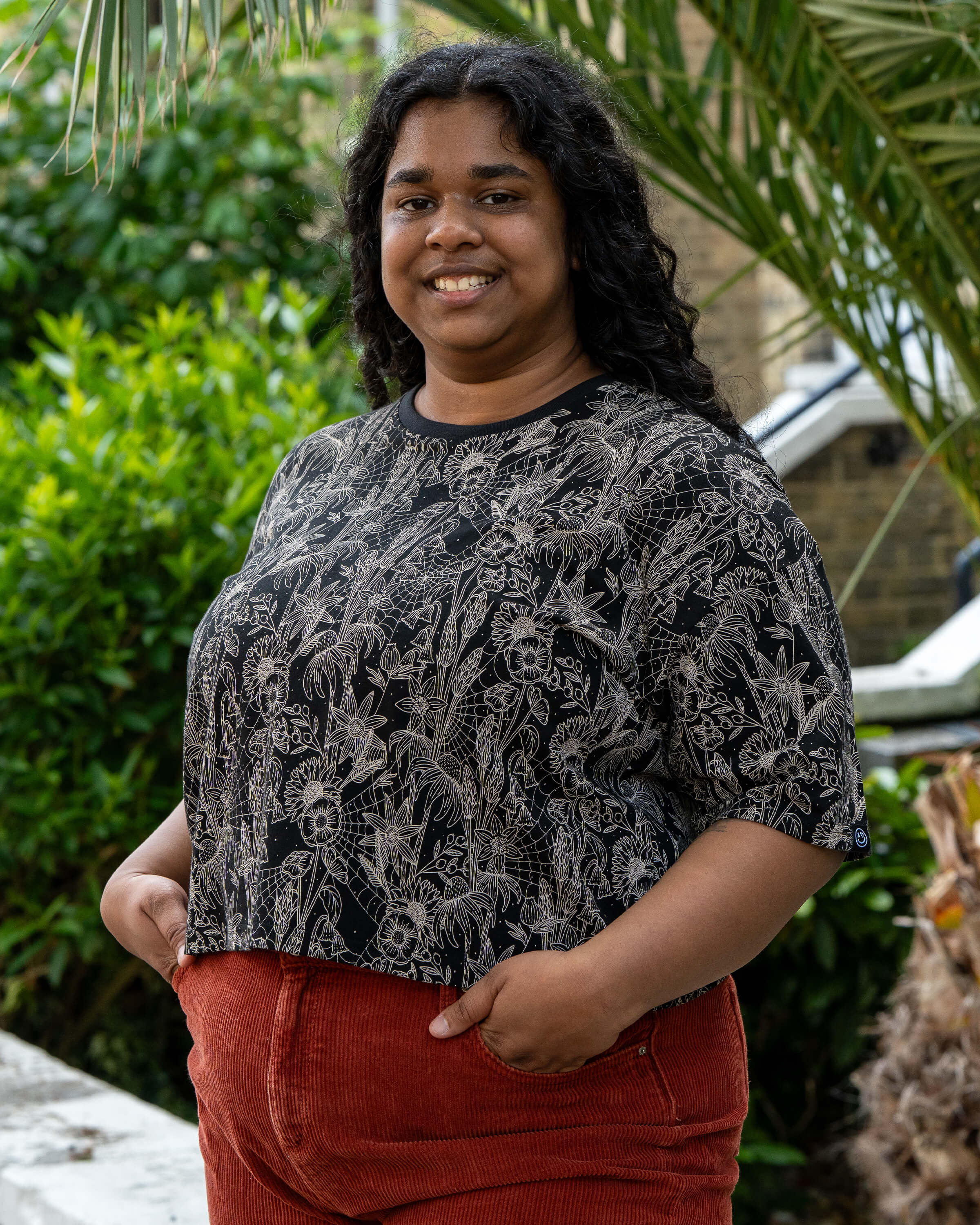 Vidhya smiles in the Rebel Romance Black Floral Cobweb Cropped Boxy Tee, hands in pockets. The bold beige cobweb and wildflower design contrasts with the black tee and rust cords. Rebel Romance is a UK brand for ethical, alternative style lovers.