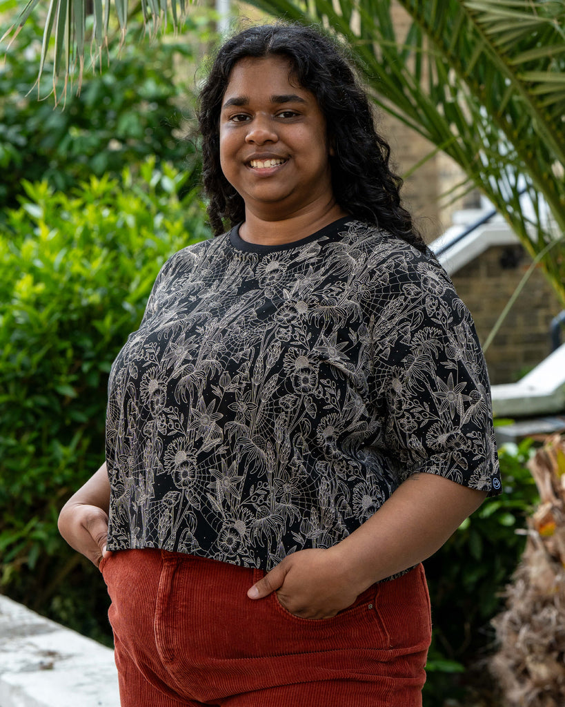 Vidhya smiles in the Rebel Romance Black Floral Cobweb Cropped Boxy Tee, hands in pockets. The bold beige cobweb and wildflower design contrasts with the black tee and rust cords. Rebel Romance is a UK brand for ethical, alternative style lovers.