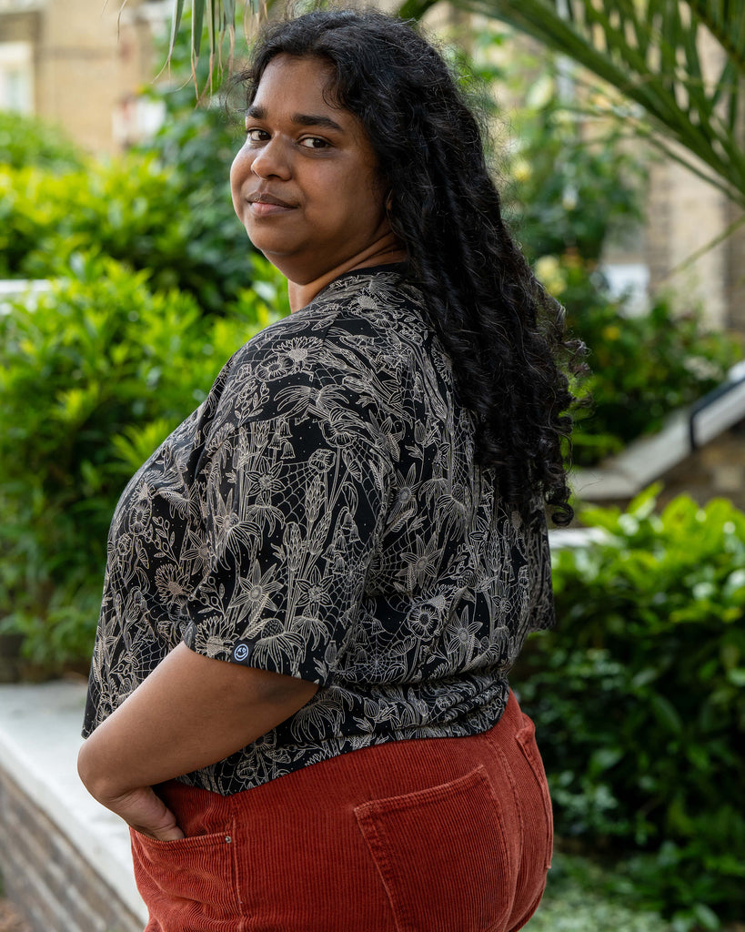 Side view of Vidhya in the Rebel Romance Black Floral Cobweb Cropped Boxy Tee. The gothic floral print stands out against the black fabric, styled with rust red cord trousers. Rebel Romance creates ethical fashion for expressive UK millennials.