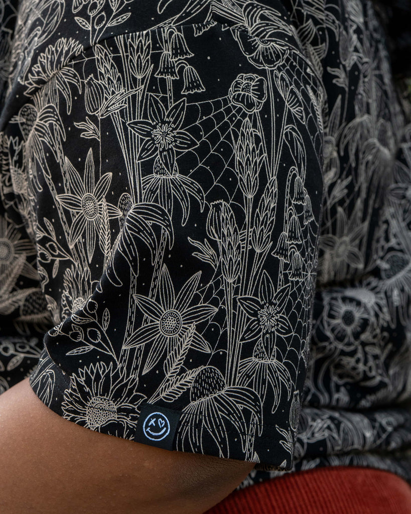 Close-up of the Rebel Romance Black Floral Cobweb Cropped Boxy Tee sleeve, highlighting the intricate beige spiderweb and floral print on black cotton. A small Rebel Romance logo tag is visible. Designed by a UK indie brand celebrating bold, alternative fashion.