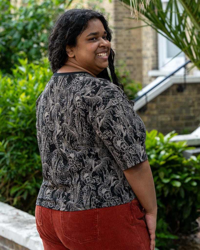 Vidhya models the Rebel Romance Black Floral Cobweb Cropped Boxy Tee from the back, smiling over her shoulder. The black top features an all-over beige floral and spiderweb print. Styled with rust corduroy trousers. Rebel Romance is a UK brand championing alternative fashion for millennials.