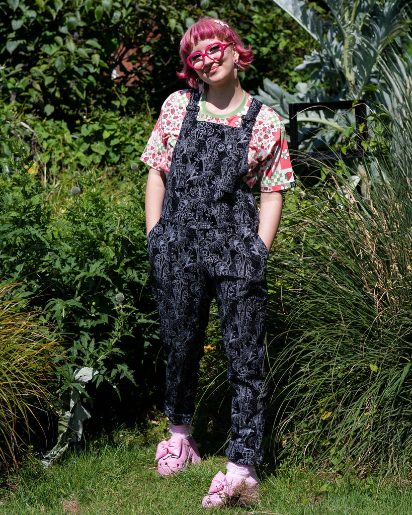Jess, a person with short pink bobbed hair wearing the rebel romance strawberry picnic crop tee with the black floral cobweb dungarees and pink heeled crocs. She is stood outside in a park smiling to camera with her hands in the side pockets. The dungaree print is a black base with white floral outlines, cobwebs and sparkles. 