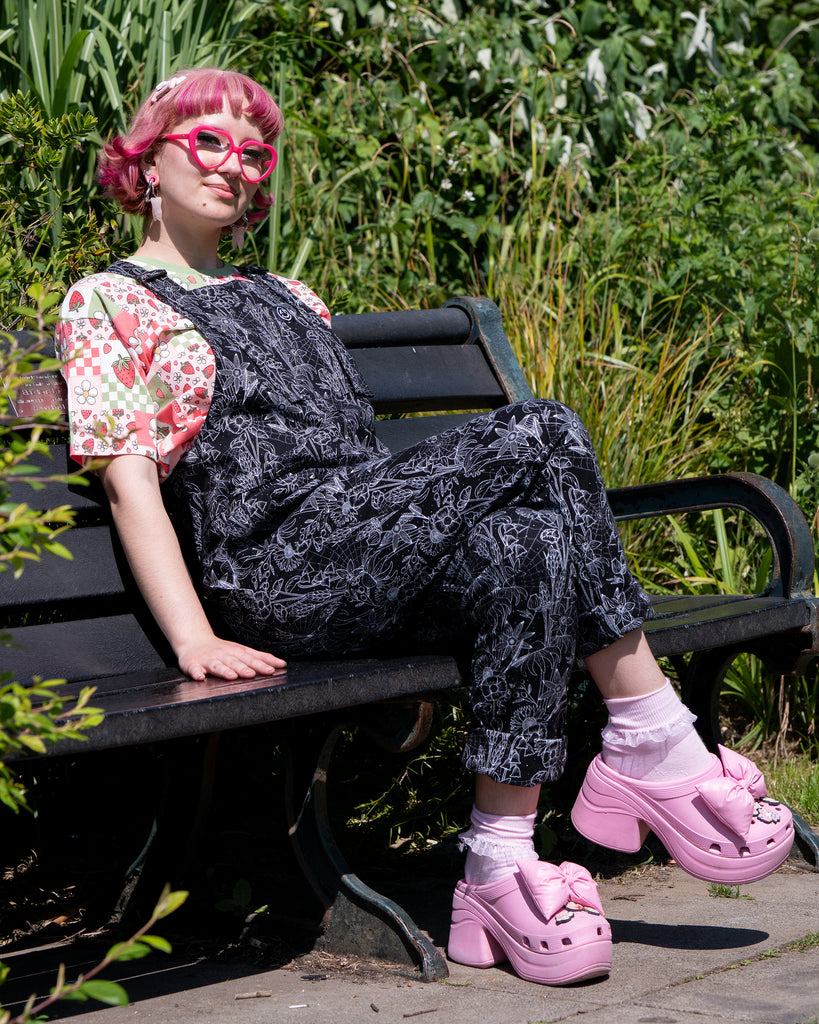 Jess, a person with short pink bobbed hair, sparkly pink makeup and heart glasses wearing the rebel romance strawberry picnic crop tee with the black floral cobweb dungarees. She is sat outside on a park bench smiling to camera. The dungaree print is a black base with white floral outlines, cobwebs and sparkles. 