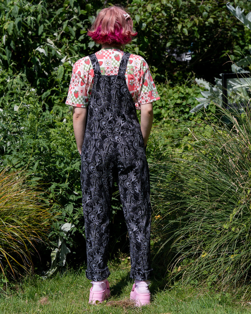 Jess, a person with short pink bobbed hair wearing the rebel romance strawberry picnic crop tee with the black floral cobweb dungarees. She is stood outside in a park facing away with her hands in the pockets to show the back details. The dungaree print is a black base with white floral outlines, cobwebs and sparkles. 