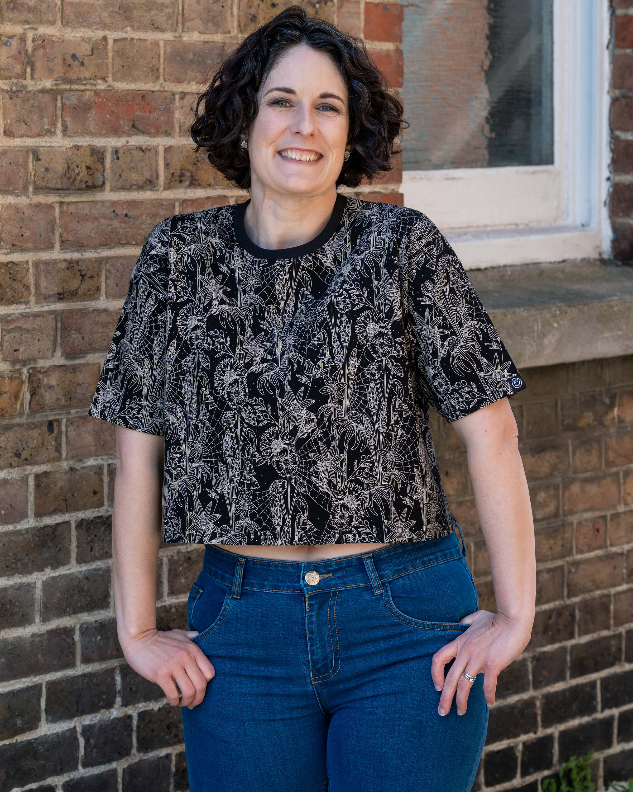 Sarah poses in front of a brick wall, smiling in the Rebel Romance Black Floral Cobweb Cropped Boxy Tee. The intricate floral and cobweb print pops against the black fabric. Paired with bright blue jeans. Rebel Romance is a UK small business celebrating alternative style and self-expression.