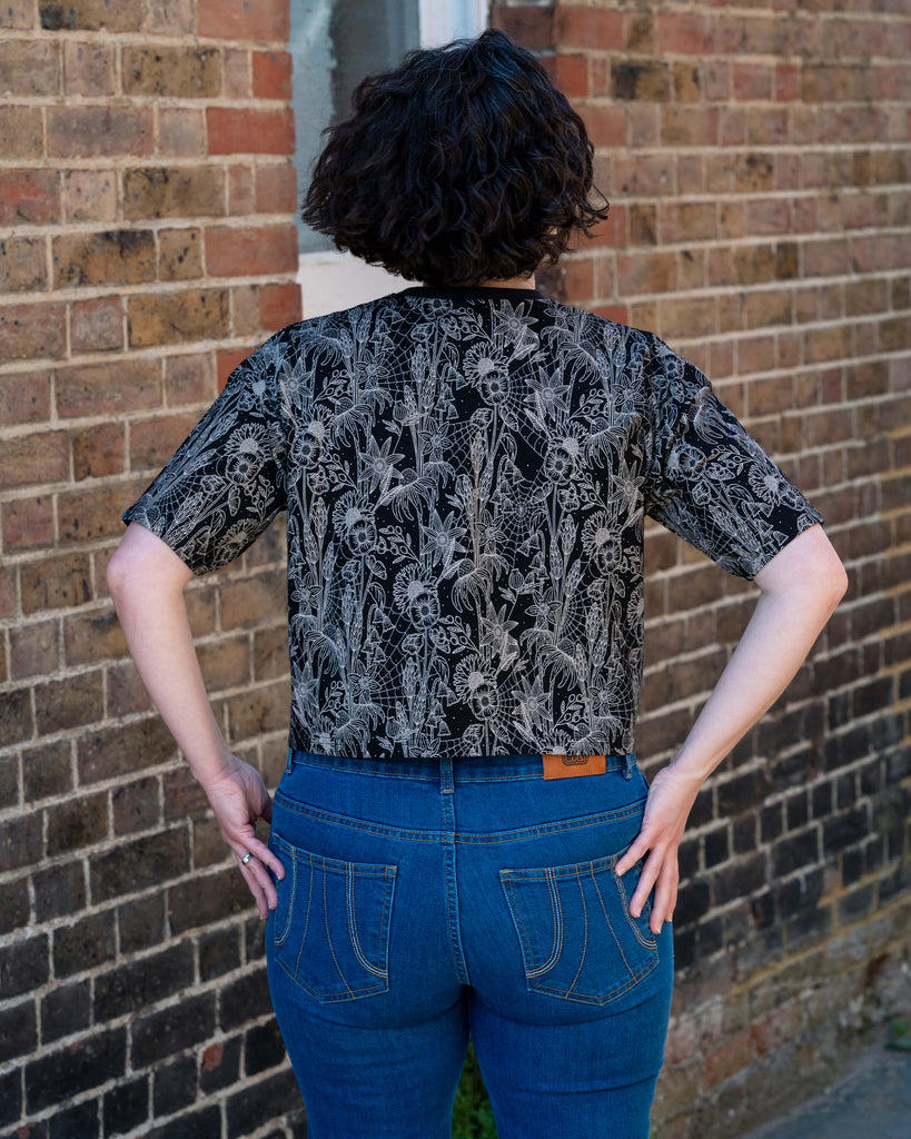 Back view of Sarah modelling the Rebel Romance Black Floral Cobweb Cropped Boxy Tee. The gothic floral and spiderweb pattern stands out against the black fabric. Paired with blue denim jeans. Rebel Romance offers ethical and expressive fashion for UK millennials.