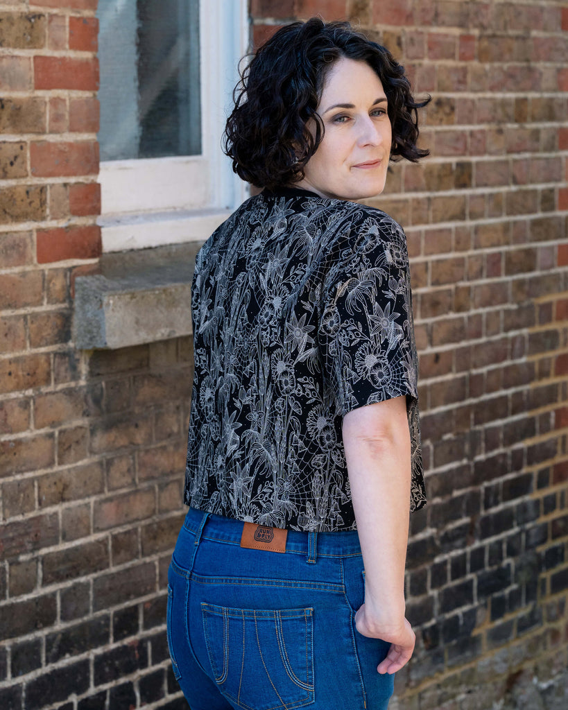 Sarah wears the Rebel Romance Black Floral Cobweb Cropped Boxy Tee, turning to look over her shoulder. The boxy crop top features a beige gothic floral and cobweb print on black. Styled with high-waisted blue jeans. Rebel Romance is a UK brand celebrating alternative style for bold millennials.