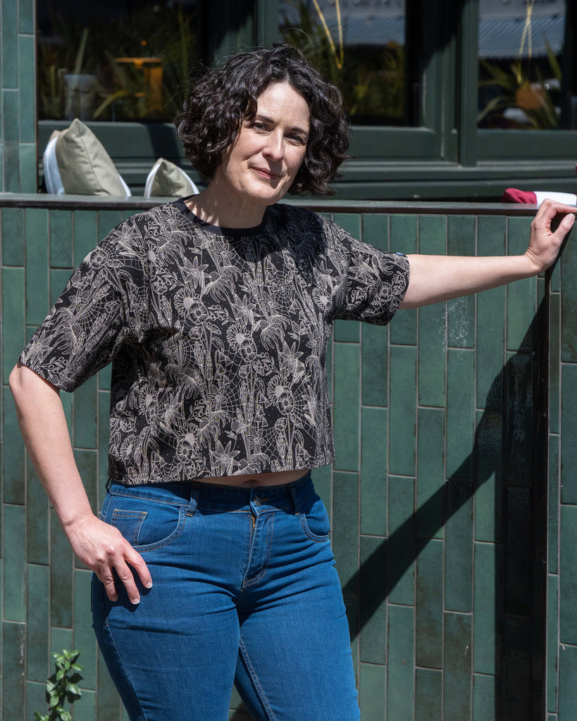 Sarah models the Rebel Romance Black Floral Cobweb Cropped Boxy Tee outdoors, resting her arm against green tiles. The crop top features a beige floral and spiderweb pattern over black. Styled with blue skinny jeans. Rebel Romance champions bold, millennial-friendly fashion in the UK.