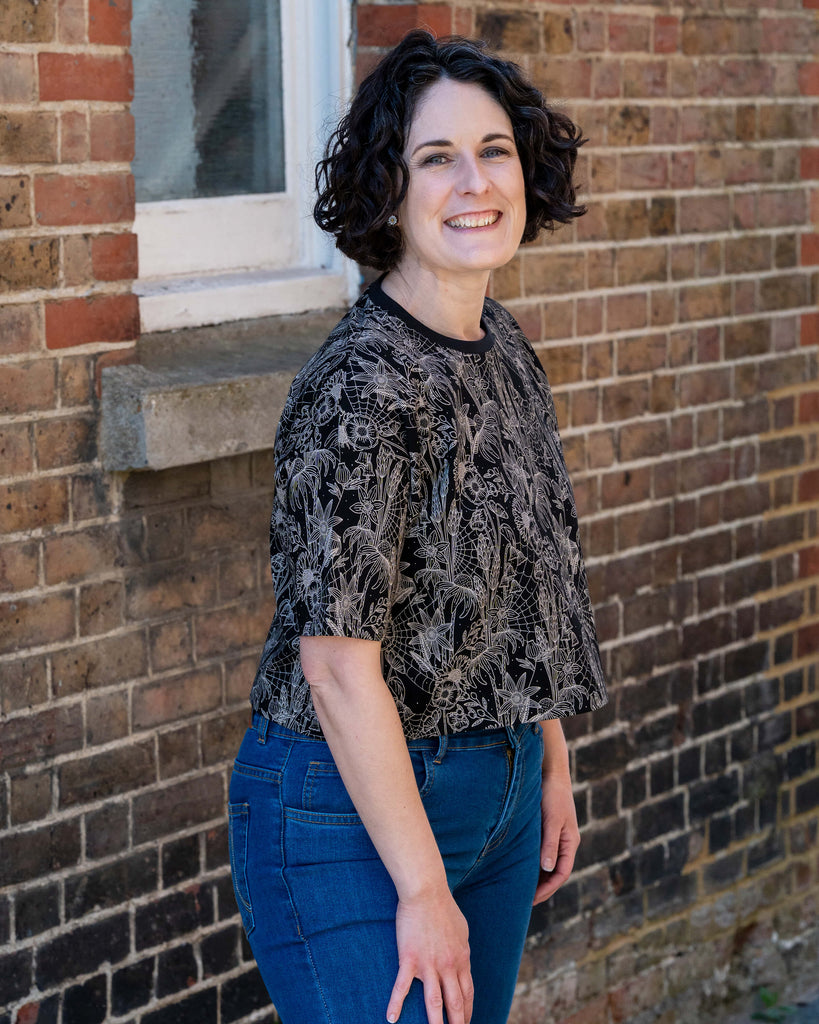 Sarah smiles in the Rebel Romance Black Floral Cobweb Cropped Boxy Tee. The tee features a striking floral and cobweb print in beige on black, styled with high-rise blue jeans. Rebel Romance celebrates alternative UK fashion with slow, ethical flair.