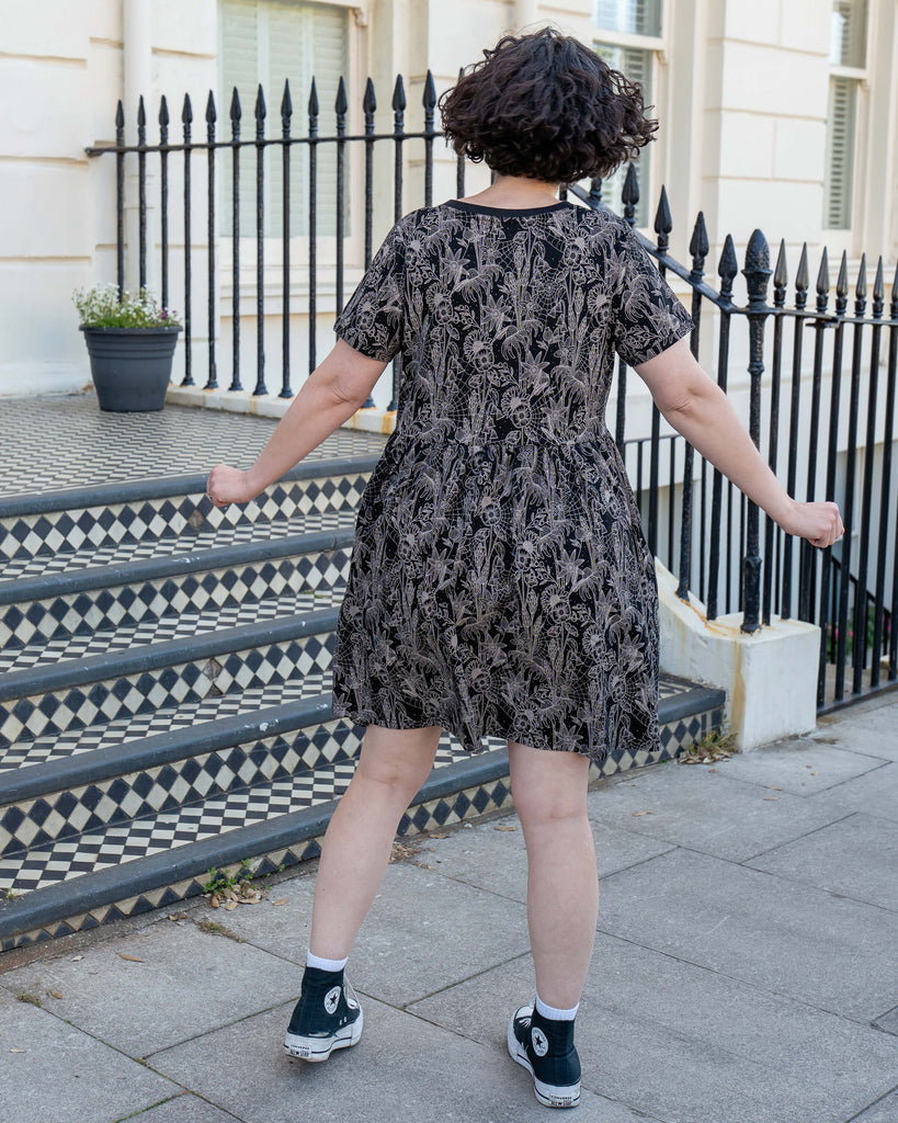 Back view of Sarah twirling outside a Brighton townhouse in the Rebel Romance Black Floral Cobweb Stretch Smock Dress. The bold monochrome floral and web print adds witchy whimsy to this comfy, size-inclusive silhouette, made for everyday magic.