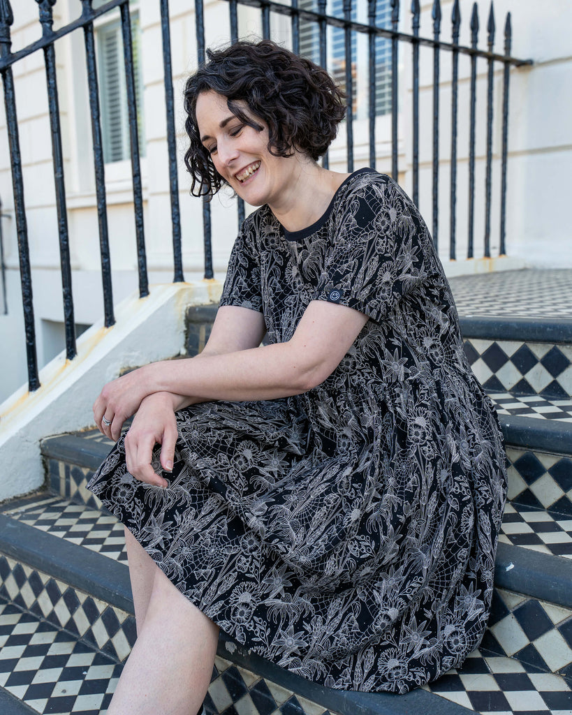 Sarah laughs looking down and sits on patterned steps in the Rebel Romance Black Floral Cobweb Stretch Smock Dress. This short-sleeved dress blends gothic charm with botanical illustration, making it a statement piece for millennial dopamine dressers who love slow fashion.
