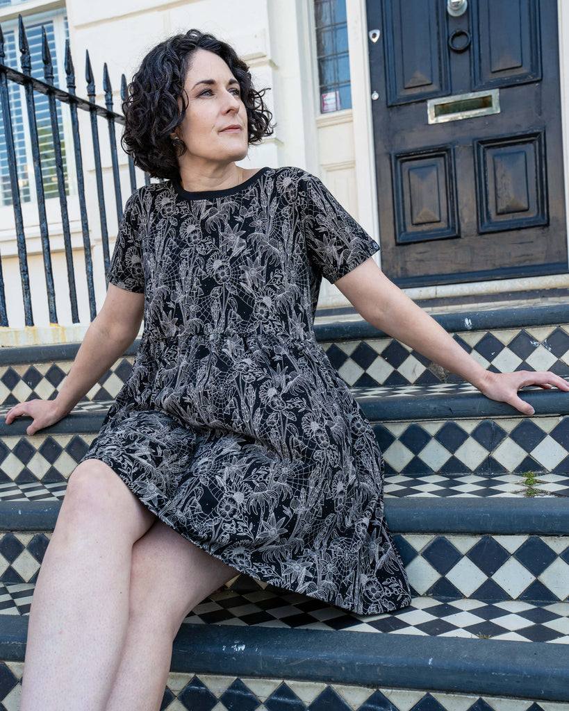 Sarah sits confidently on Victorian-style black and white tiled steps wearing the Rebel Romance Black Floral Cobweb Stretch Smock Dress. This comfy, gothic-inspired dress features intricate floral and spiderweb linework—perfect for alternative fashion lovers seeking ethical style with edge.
