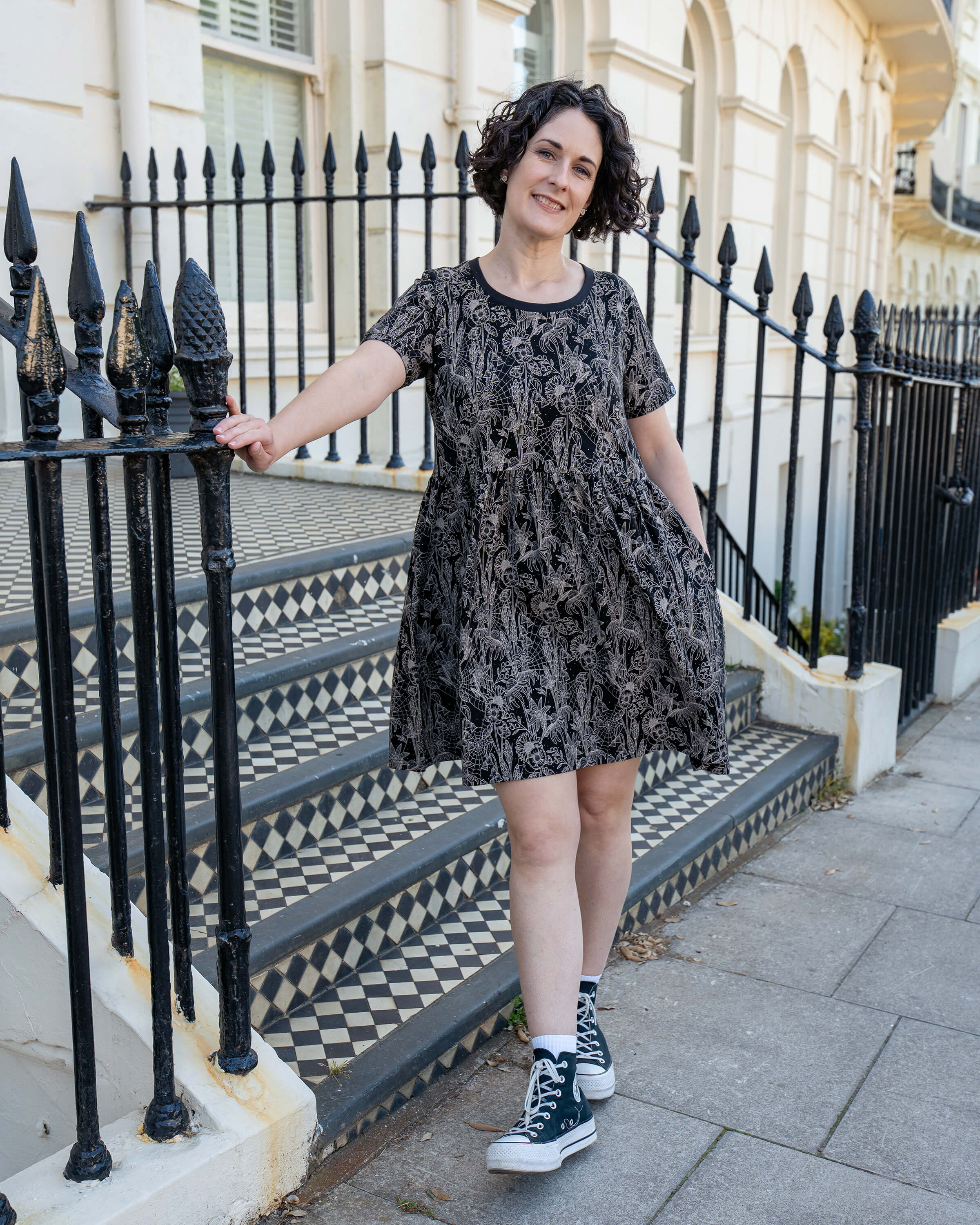 Sarah poses with one hand on a rail, wearing the Rebel Romance Black Floral Cobweb Stretch Smock Dress. This ethical, alternative fashion staple flatters with a soft gathered waist, short sleeves, and magical monochrome patterning.