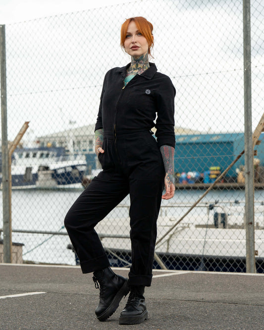 Erin stands outdoors in front of a fence by the docks wearing a black stretch twill boiler suit with a zip front, hands in pockets, and chunky black boots.