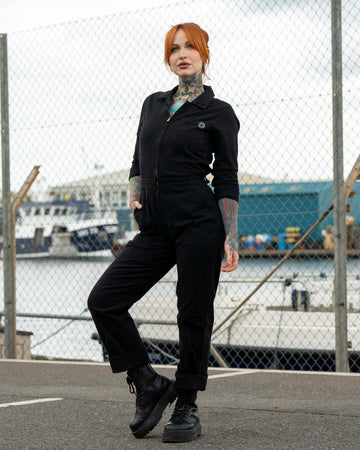 Erin stands outdoors in front of a fence by the docks wearing a black stretch twill boiler suit with a zip front, hands in pockets, and chunky black boots.