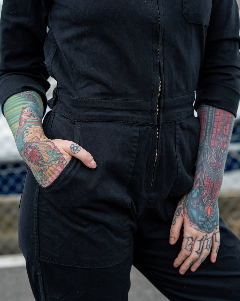 Close-up detail of the boiler suit waist, showing Erin’s tattooed hands resting in the deep side pockets with visible front zip.