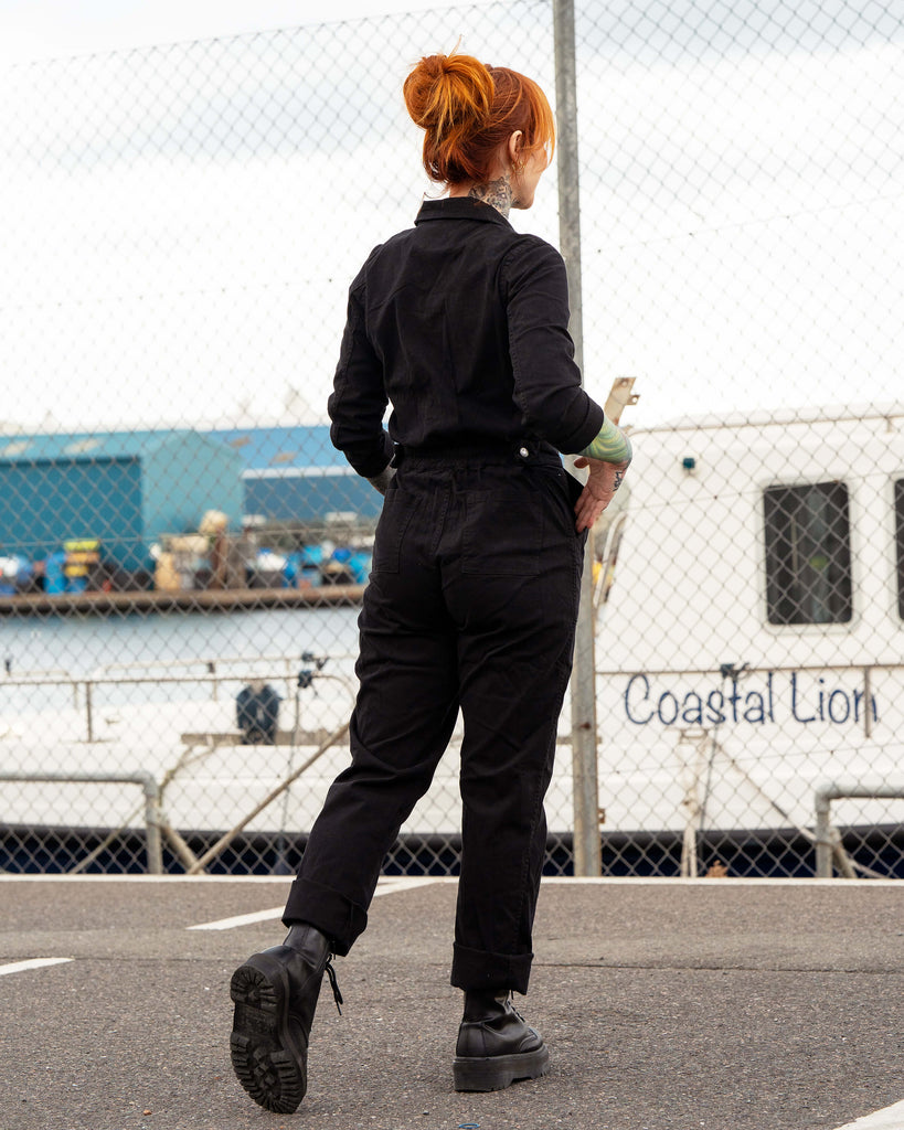 Erin faces away from the camera, walking near the dockside in the black boiler suit with turned-up cuffs and black boots.