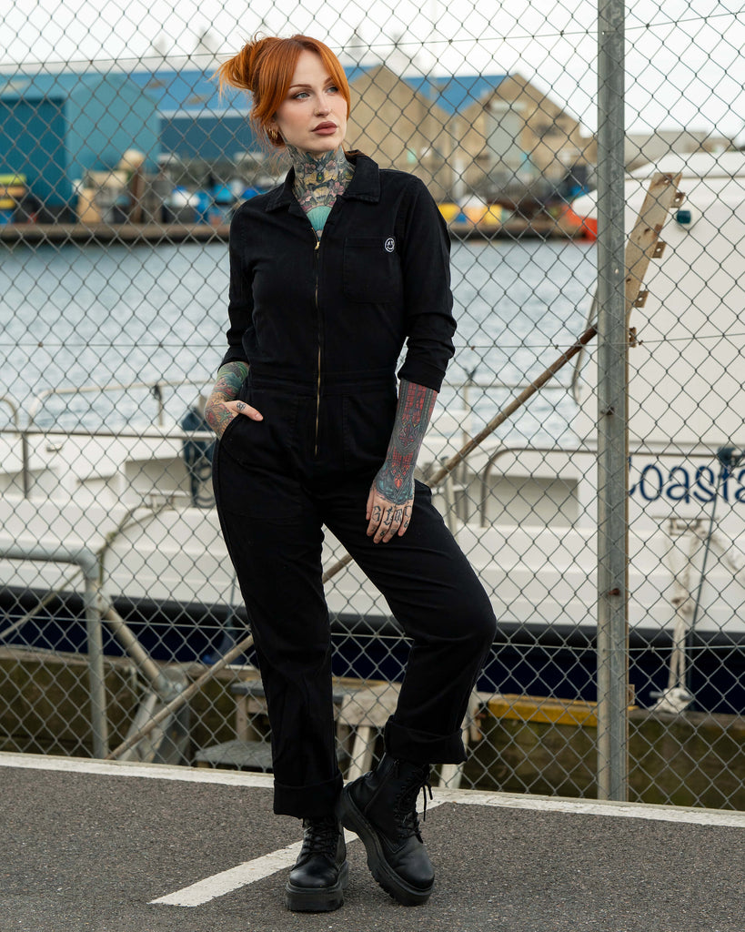 Erin stands relaxed in the black stretch twill boiler suit with hands in pockets, posed in front of docked boats and chain-link fencing.