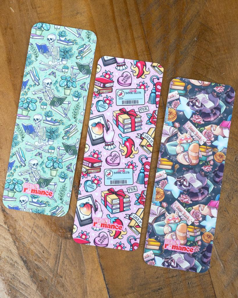 A flat lay of three colourful bookmarks on a wooden surface. Each design has a different aesthetic: skeleton and plants, romantic book club, and Smut Trash with raccoons and red pandas.
