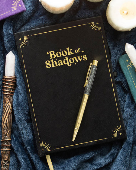 Flat lay of the Book of Shadows journal and pen, styled with candles, crystals, and wands on plush dark fabric. The journal's gold detailing and black velvet cover create a mystical vibe.