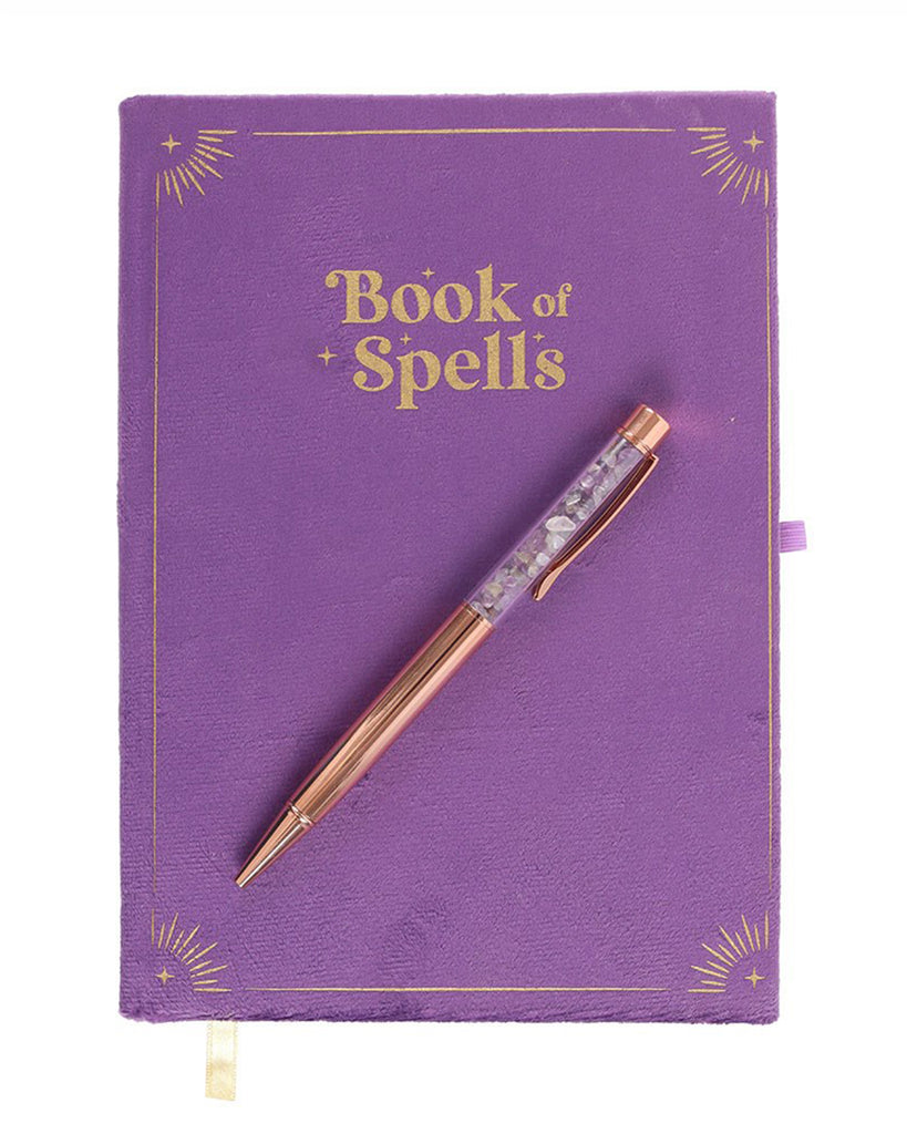 Flat lay of the Book of Spells journal with an amethyst crystal pen. The purple velvet cover features gold-foiled sunburst corner details and the title Book of Spells in a mystical, serif font.