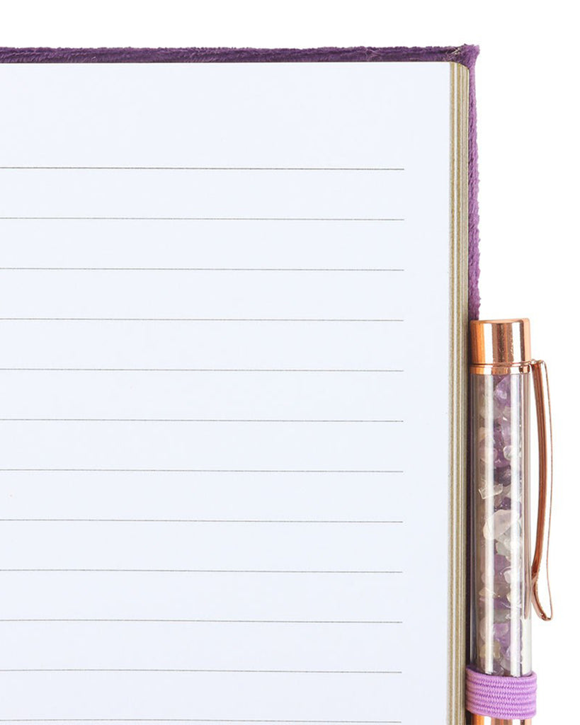 Interior view of the Book of Spells journal, showing lined white pages with a gold trim. The amethyst crystal pen rests in a purple elastic holder, matching the journal's plush cover.