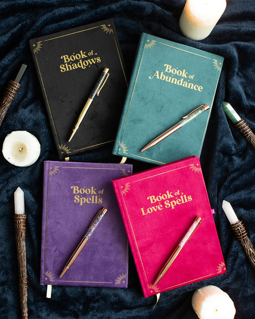 Flat lay of four plush velvet journals from Rebel Romance on a dark, soft fabric background. Titles include Book of Shadows, Book of Abundance, Book of Spells, and Book of Love Spells, each paired with a matching crystal pen. The Book of Spells is purple with gold detailing and an amethyst crystal pen for clarity and mindfulness.