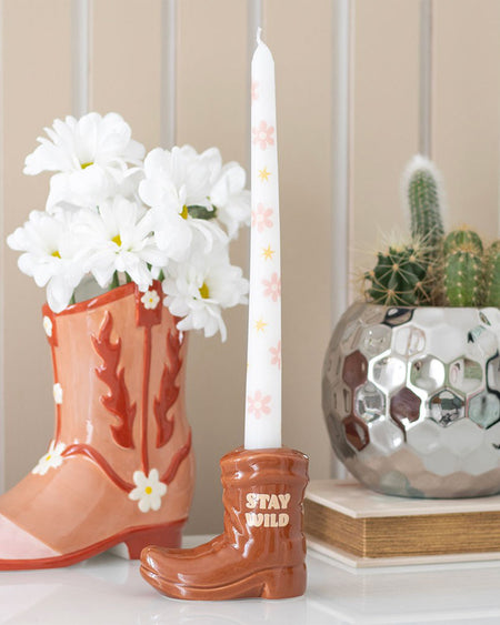 Styled shot of the Rebel Romance brown cowboy boot candle holder on a white shelf, surrounded by daisies, a cactus, and other western-inspired decor. The playful "Stay Wild" slogan and retro floral taper candle add a quirky, alternative aesthetic.