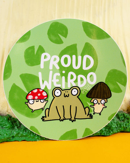 A round green vinyl sticker by Katie Abey, sold at Rebel Romance, featuring the words “Proud Weirdo” in white bold text. The design includes a cartoon frog with a happy expression, flanked by two quirky mushrooms—one red with white spots, the other brown. Surrounded by lily pad shapes, this sticker celebrates individuality and joyful weirdness.