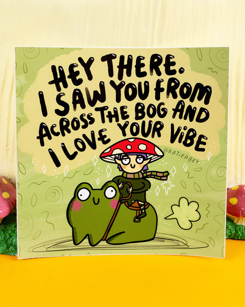 A square green vinyl sticker by Katie Abey, sold by Rebel Romance, featuring a cheerful cartoon frog with pink cheeks being ridden by a smiling mushroom elf. Above them is bold black text that reads: “Hey there. I saw you from across the bog and I love your vibe.” A whimsical, playful design perfect for celebrating weirdness and spreading good vibes