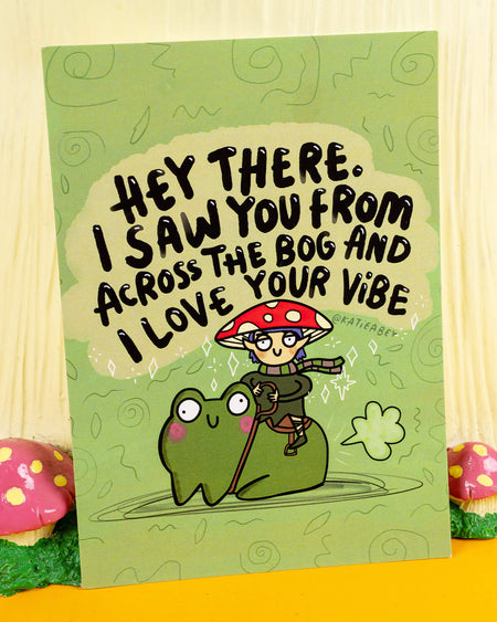 A green A6 postcard designed by Katie Abey, available at Rebel Romance, a UK small biz known for quirky, alternative vibes. The card features a mushroom elf character riding a happy cartoon frog through a bog, with the text: “Hey there. I saw you from across the bog and I love your vibe.” A playful, whimsical design perfect for spreading positive energy and flirty fun.