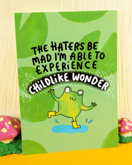 A vibrant green A6 postcard designed by Katie Abey, sold by Rebel Romance, a UK brand celebrating alternative style. The postcard features a cheerful cartoon frog in yellow wellies splashing in a puddle, with the bold text: “The haters be mad I’m able to experience childlike wonder.” Surrounded by sparkles and a playful vibe, this quirky design is perfect for spreading joy, positivity, and unapologetic froggy fun.