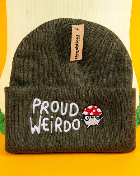 Olive green "Proud Weirdo" beanie with whimsical white text and a red-spotted mushroom character in glasses, showcasing Katie Abey's unique and quirky style. Small business Katie Abey inspires embracing your inner weirdness. Beanie displayed on a fun woodland-inspired setup.