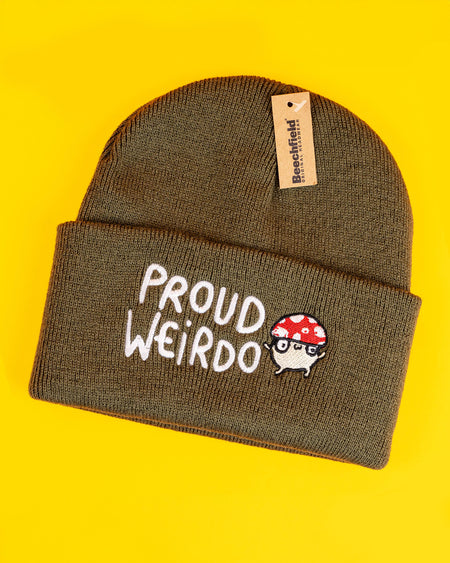 Olive green knitted beanie featuring "Proud Weirdo" text in playful white embroidery alongside an adorable mushroom character with a red spotted cap and glasses. Katie Abey's quirky, illustrated design encourages individuality and celebrates weirdness. Beanie shown flat with vibrant yellow background.