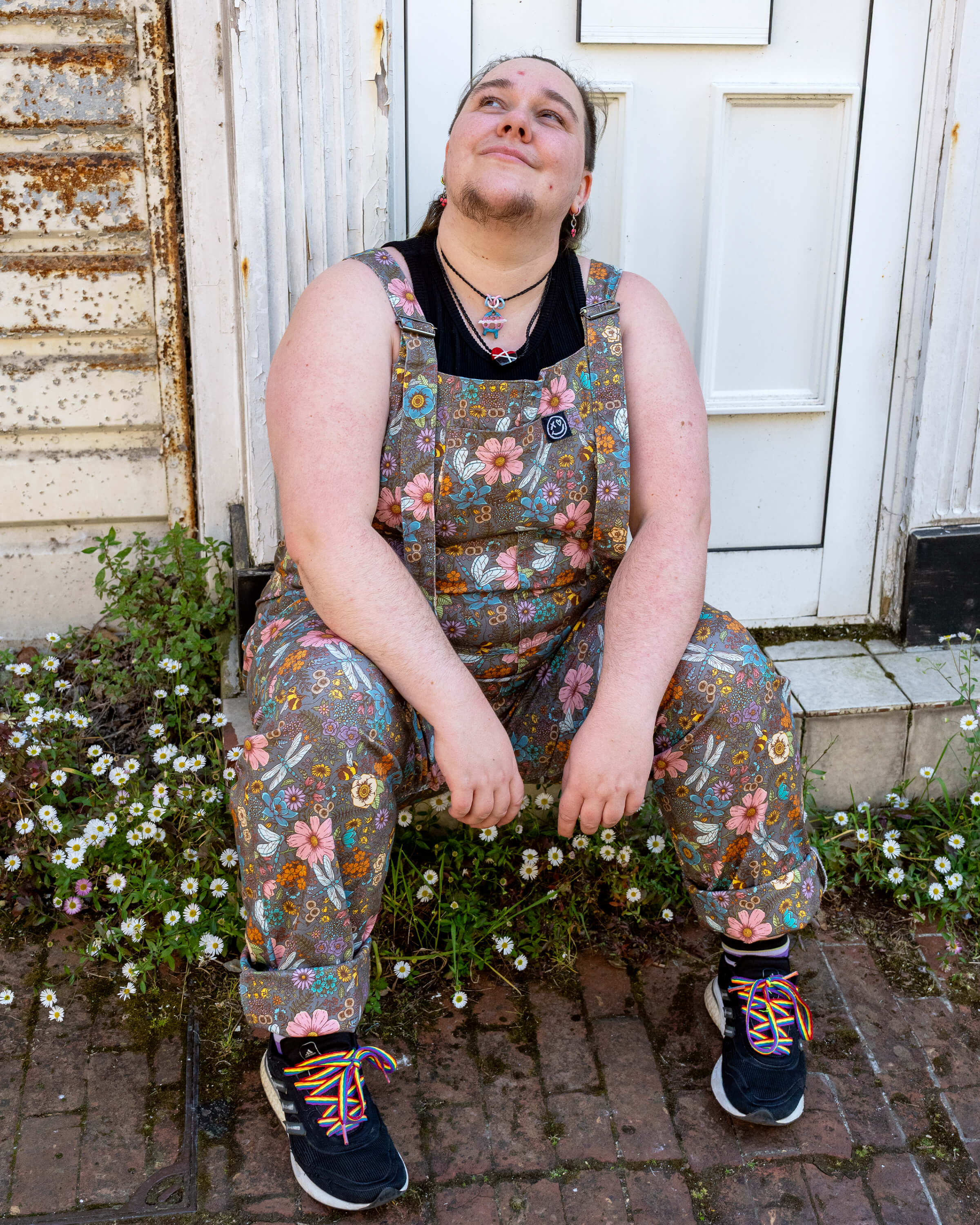 Bryony May sits in front of a white door, wearing grey stretch twill dungarees with a colourful floral and mushroom print in size 3XL. Styled with a black top and rainbow-laced trainers, they pose surrounded by daisies growing through the bricks. The look is from a small ethical slow fashion brand.