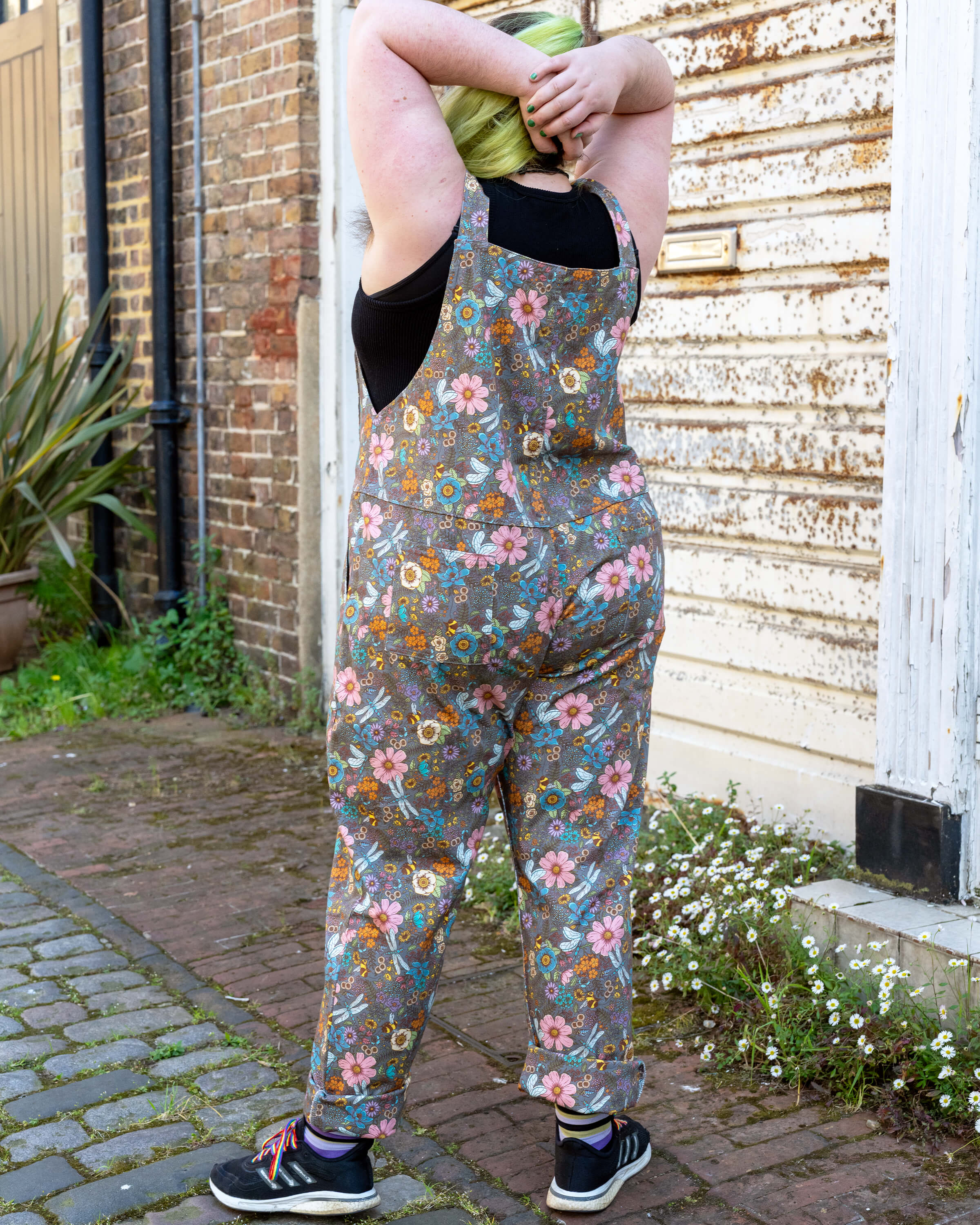 Back view of Bryony May wearing floral grey stretch twill dungarees in size 3XL, paired with a black sleeveless top and rainbow-laced trainers. They stand with arms raised behind their head, showing off the rear pockets and relaxed fit. Shot on a cobbled path with rustic walls and plants.