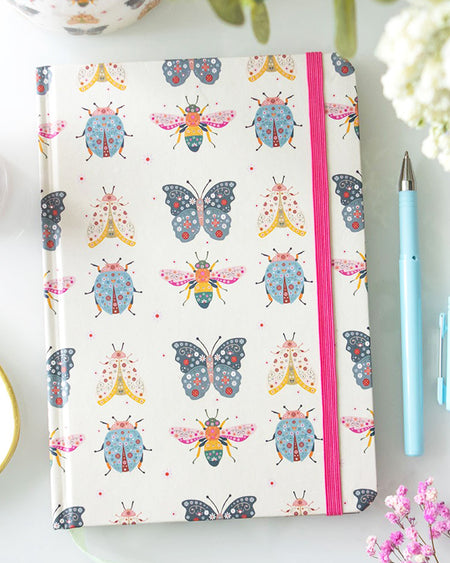 A flat lay of a cream A5 notebook with a colourful insect print featuring butterflies, bees, moths, and ladybirds. It has a bright pink elastic closure and is styled on a white desk with flowers and pastel stationery.