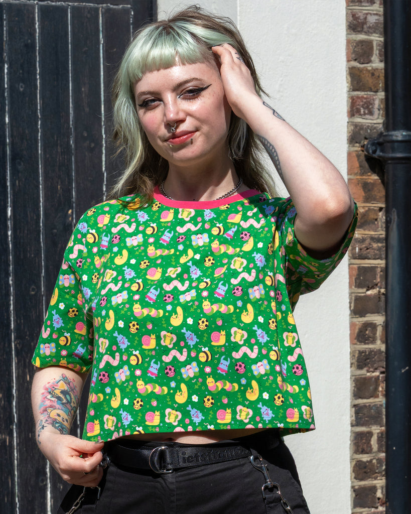 Faeryn lifts one hand to their hair and slightly lifts the Rebel Romance x Dipstick Store Bugs Cropped Boxy Tee, revealing its relaxed fit. The bug print and pink neckline stand out against black trousers. Inclusive, playful fashion from UK brand Rebel Romance.