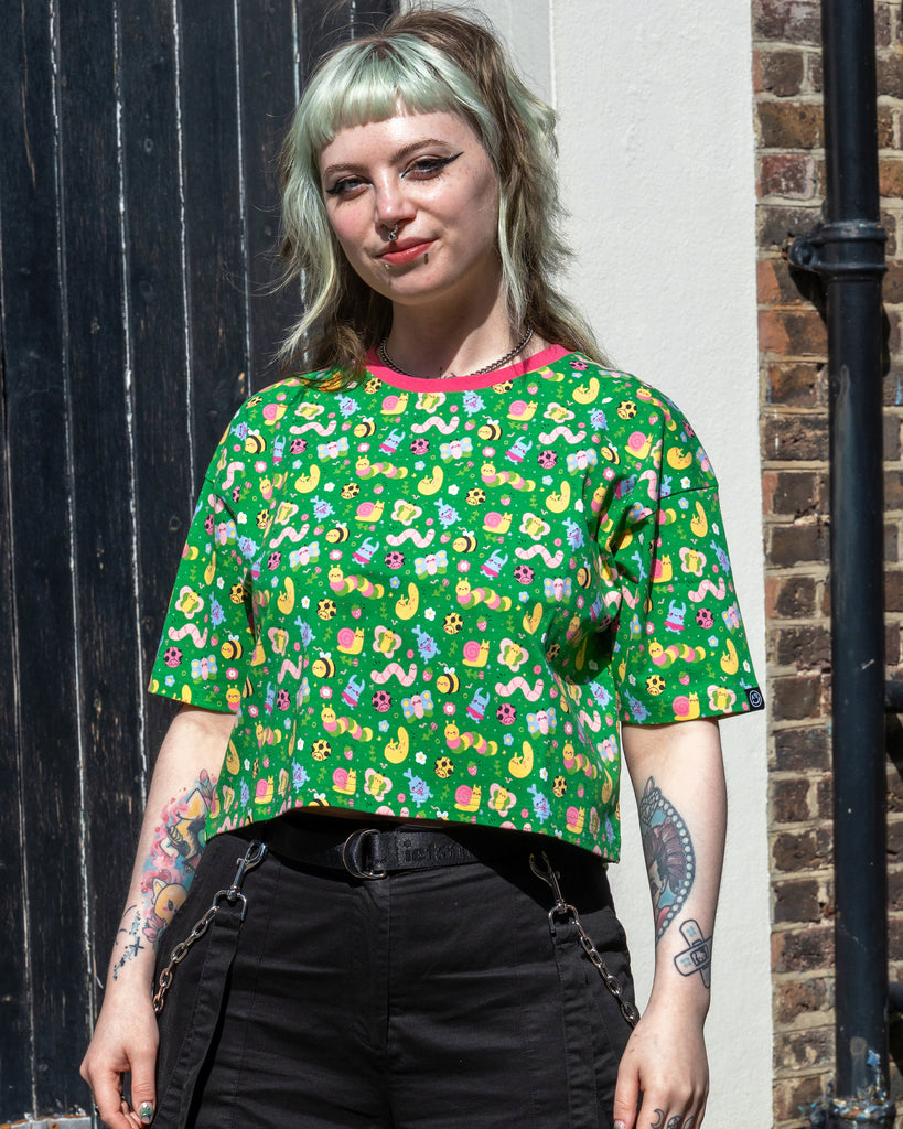 Faeryn stands confidently in the Rebel Romance x Dipstick Store Bugs Cropped Boxy Tee, smiling slightly. The cropped green top features colourful worms, butterflies and snails. Styled with black trousers and chain details. Ethical alt fashion by UK brand Rebel Romance.
