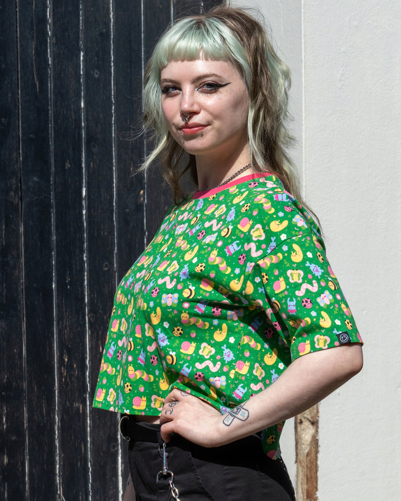 Side view of Faeryn wearing the Rebel Romance x Dipstick Store Bugs Cropped Boxy Tee with black trousers. The green crop top has a vibrant pastel bug print and a pink collar, styled with chain accessories. Inclusive slow fashion from Rebel Romance.