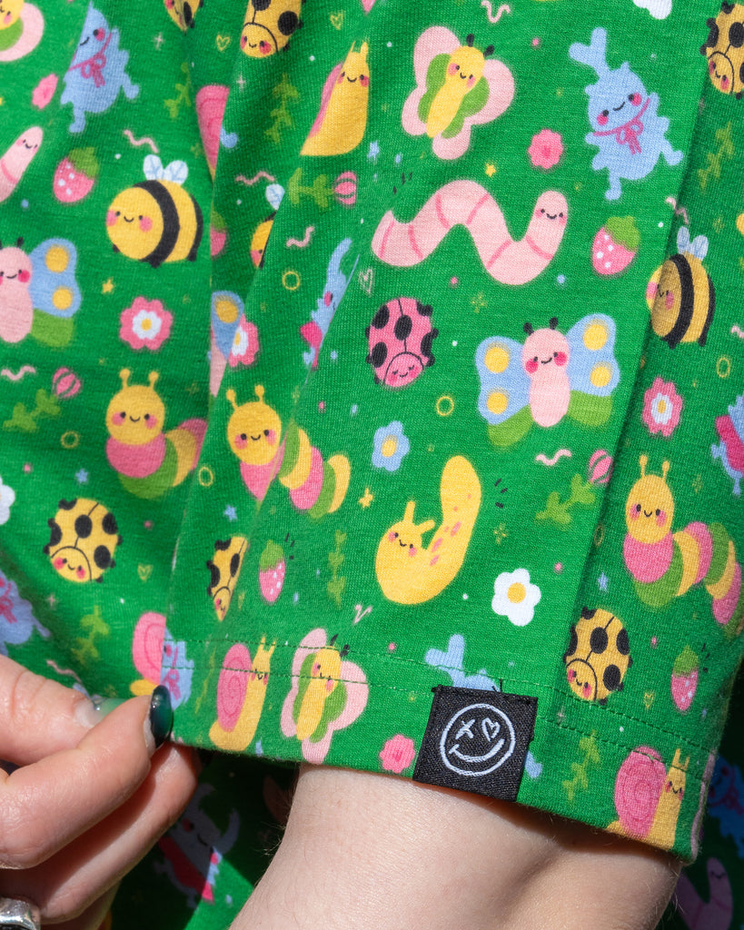 Close-up of the Rebel Romance x Dipstick Store Bugs Cropped Boxy Tee sleeve, showing the pastel bug print featuring worms, snails, bees, butterflies, and flowers on a bright green background. The black Rebel Romance smiley face logo tag is visible. Ethical dopamine fashion made in the UK.